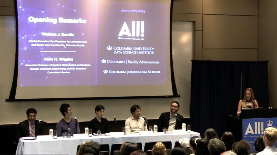 A table with a diverse group of five panelists in a full auditorium for an event.